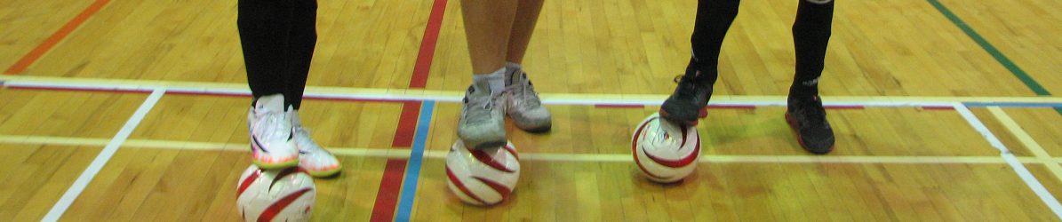Photo des jambes de 3 joueurs avec 3 ballons de cécifoot dans un gymnase avec un plancher en bois.