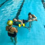 Du sport pour moi! - Natation. Montréal - Printemps 2019. Alexandra observe Marc-Antoine qui nage sur le côté avec l'aide de flotteurs aux mains et Raphaëlle pour les jambes.